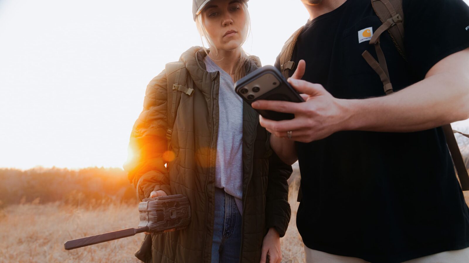 Two hunters checking trail camera images on a phone in a field at sunset